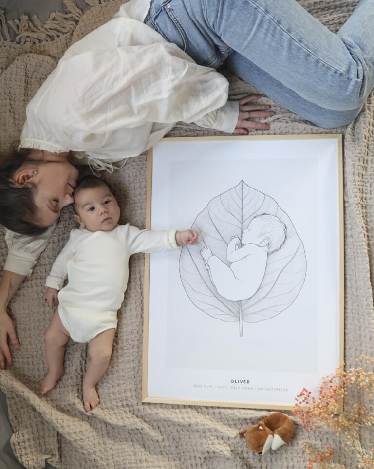 En mamma och hennes bebis ligger sida vid sida på en mjuk, beige filt. Bebisen sträcker sin hand mot en personlig födelsetavla med motivet "Baby leaf" från Stories by Mia, en illustration av ett spädbarn liggandes på ett stort blad, som visar namn, födelsedatum, tid, vikt och längd. Bredvid tavlan ligger en liten mjukdjursräv och torkade blommor – en harmonisk scen som förmedlar värme, kärlek och personligt minne.