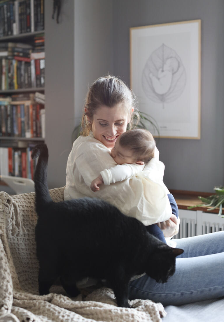 En mamma sitter i en fåtölj och håller sitt sovande barn i famnen, medan en svart katt går förbi vid sidan. I bakgrunden syns en personlig födelsetavla med motivet "Baby Leaf" från Stories by Mia. En illustration av barnet på ett stort blad, samt en bokhylla fylld med böcker. Bilden utstrålar en varm och hemtrevlig atmosfär med både närhet och familjär vardag.