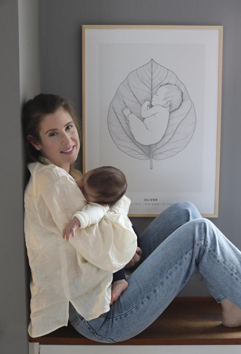 En mamma sitter lutad mot en vägg och håller sitt sovande barn i famnen framför en personlig födelsetavla från Stories by Mia. Tavlan som har motivet "Baby Leaf" visar en illustration av en nyfödd bebis liggandes på ett stort blad, med barnets namn, födelsedatum, tid, vikt och längd angivet nedtill. En varm och kärleksfull scen som förmedlar närhet och minnen.